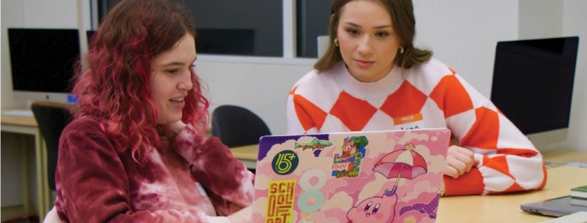 Two student, Anna McCreary and Trinity Czarnik gather in front of a laptop in the design studio.