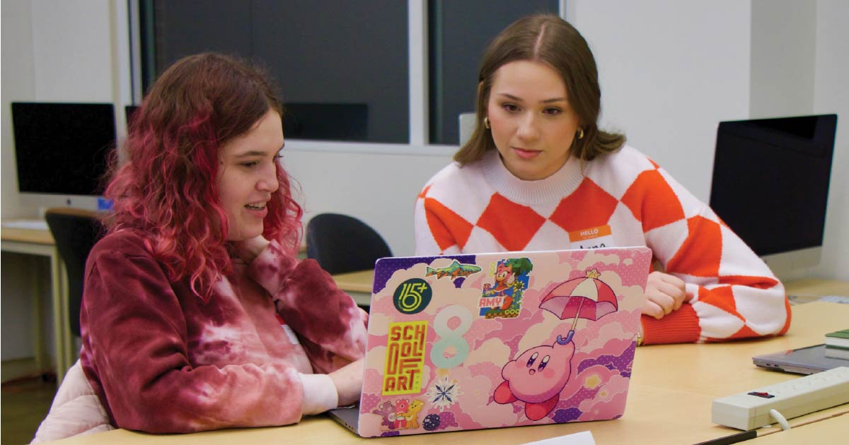 Two student, Anna McCreary and Trinity Czarnik gather in front of a laptop in the design studio.