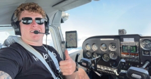 Luke Dalton sits in a cockpit in sunglasses taking a selfie with a thumbs up.