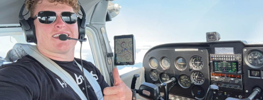Luke Dalton sits in a cockpit in sunglasses taking a selfie with a thumbs up.