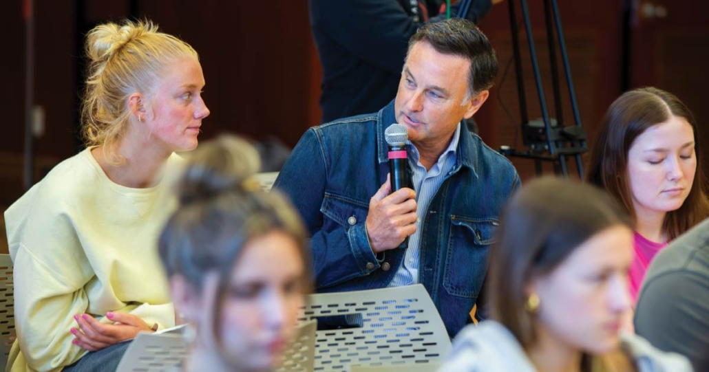 Mike sits next to a student holding a microphone.