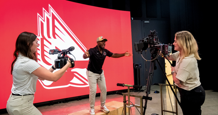 Two students filming a man standing in front of the new LED volume wall that's displaying the athletics cardinal logo