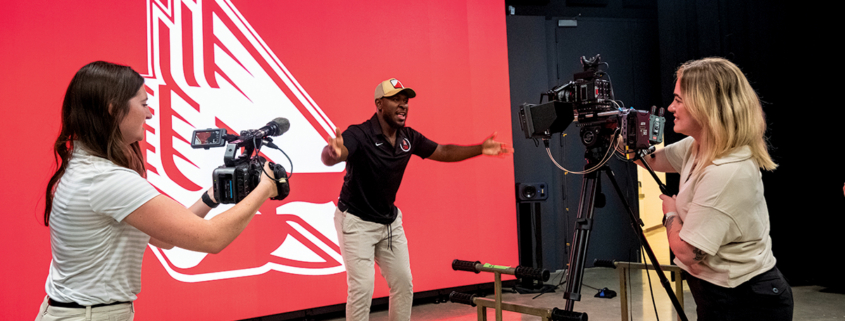 Two students filming a man standing in front of the new LED volume wall that's displaying the athletics cardinal logo