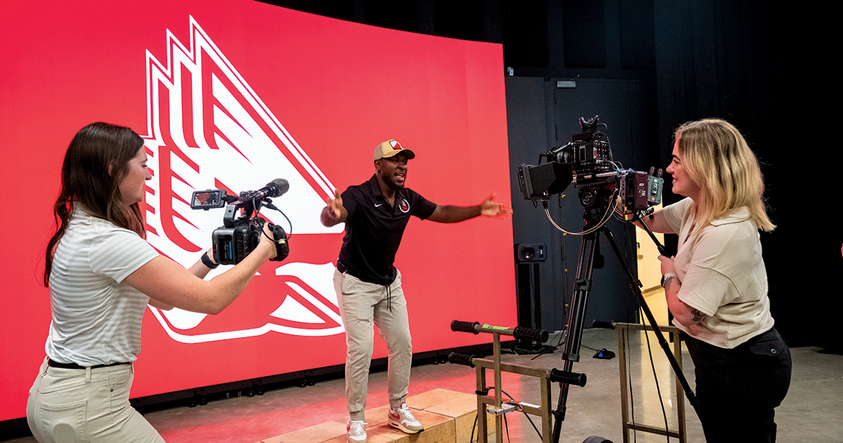 Two students filming a man standing in front of the new LED volume wall that's displaying the athletics cardinal logo