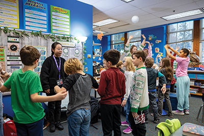 Elementary students stand in a classroom participating in an activity led by a teacher.