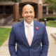 Smiling man (Kieth A. Robinson) in a blue suit and pink tie poses outdoors in front of a brick building
