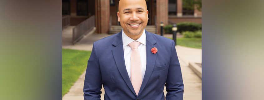 Smiling man (Kieth A. Robinson) in a blue suit and pink tie poses outdoors in front of a brick building