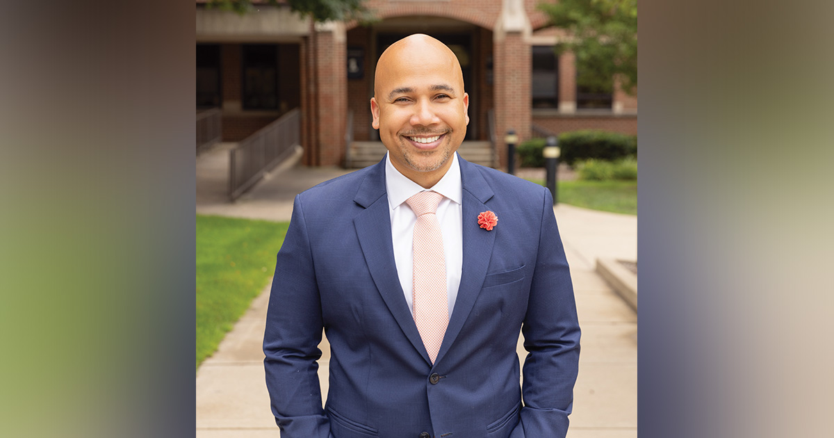 Smiling man (Kieth A. Robinson) in a blue suit and pink tie poses outdoors in front of a brick building