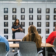 Person speaking to a small group seated at tables in a conference room with a wall of framed portraits behind them.