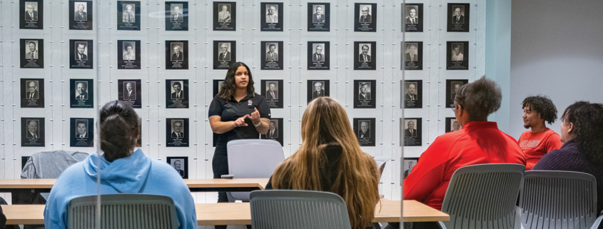 Person speaking to a small group seated at tables in a conference room with a wall of framed portraits behind them.