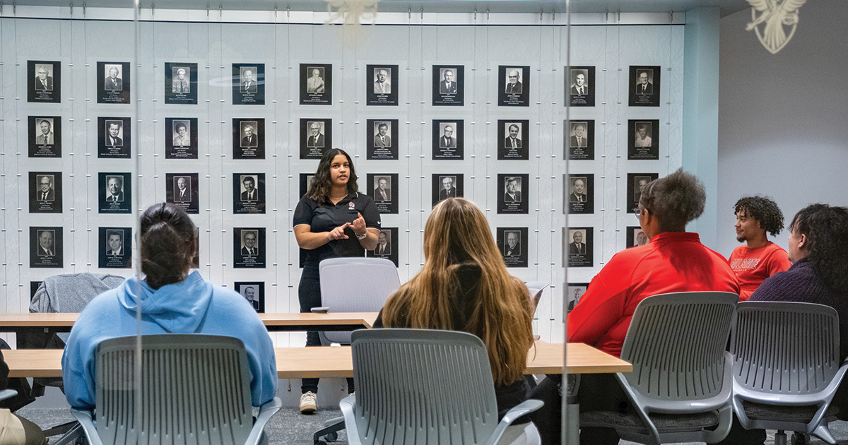 Person speaking to a small group seated at tables in a conference room with a wall of framed portraits behind them.