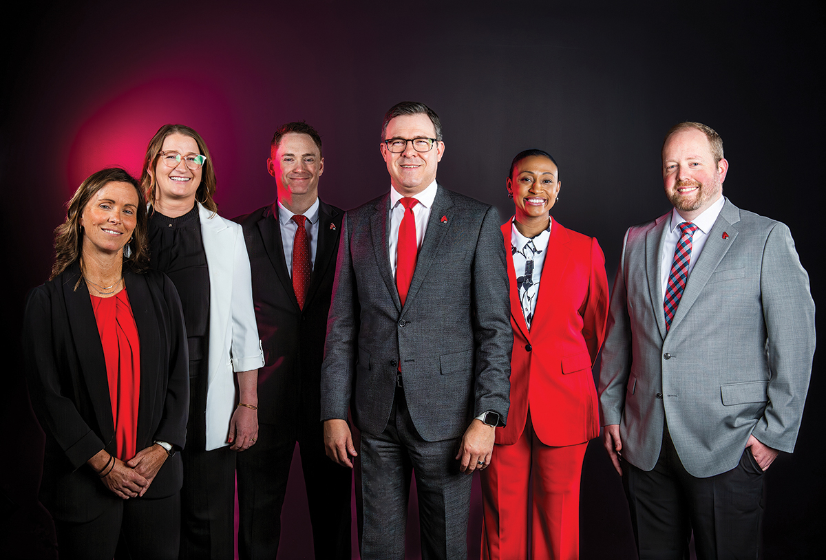 Six Ball State staff members in business attire pose together against a dark background with red back lighting.