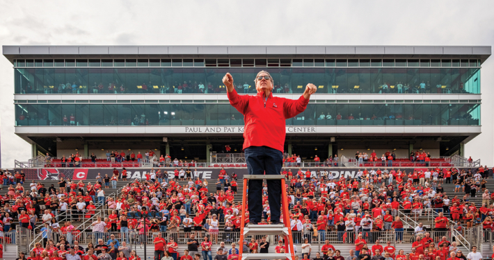 A man wearing a red long sleeved shirt stands and conducts a band from a podium in front of filled stadium bleachers.