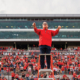 A man wearing a red long sleeved shirt stands and conducts a band from a podium in front of filled stadium bleachers.