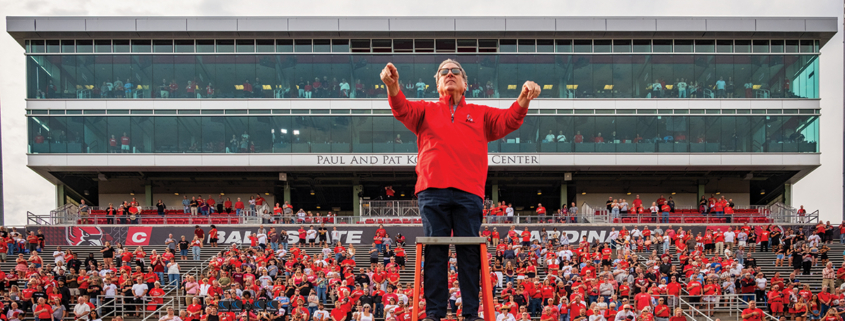 A man wearing a red long sleeved shirt stands and conducts a band from a podium in front of filled stadium bleachers.