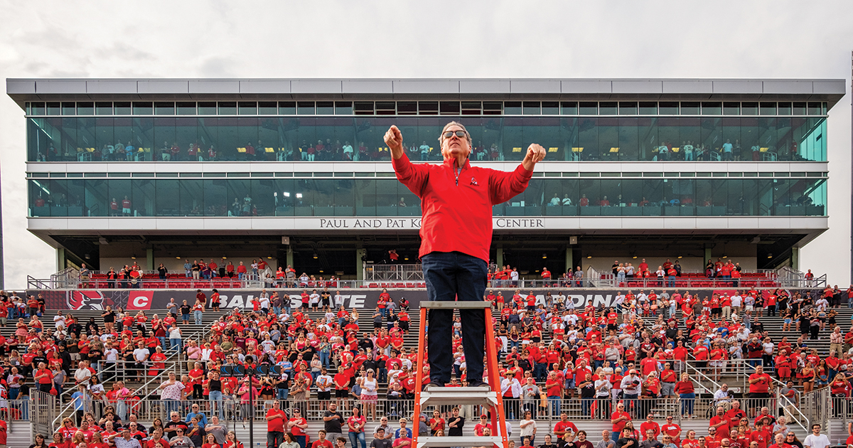 A man wearing a red long sleeved shirt stands and conducts a band from a podium in front of filled stadium bleachers.