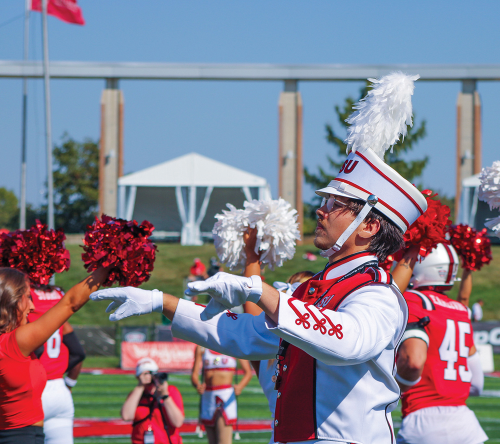 A drum major stands in uniform and conducts, Ball State cheerleaders and football players in the background.