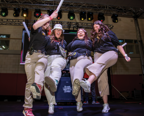 Four women are jumping together, one holds a glow stick in the air.