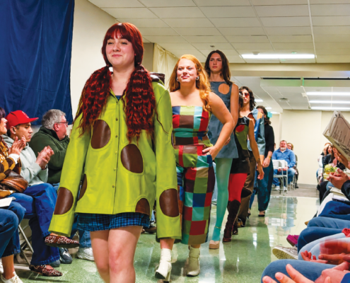People walking down a runway. A woman wears a green coat with. brown circles on it.