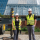 Two construction staff members wearing hard hats and reflective safety vests stand in front of a building under renovation, with construction equipment and scaffolding behind them.