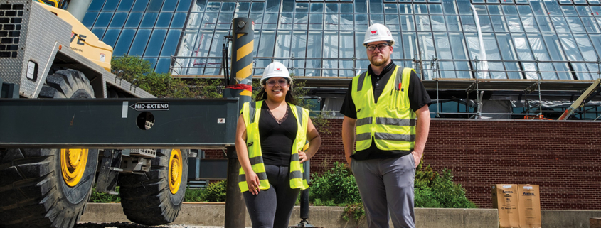 Two construction staff members wearing hard hats and reflective safety vests stand in front of a building under renovation, with construction equipment and scaffolding behind them.