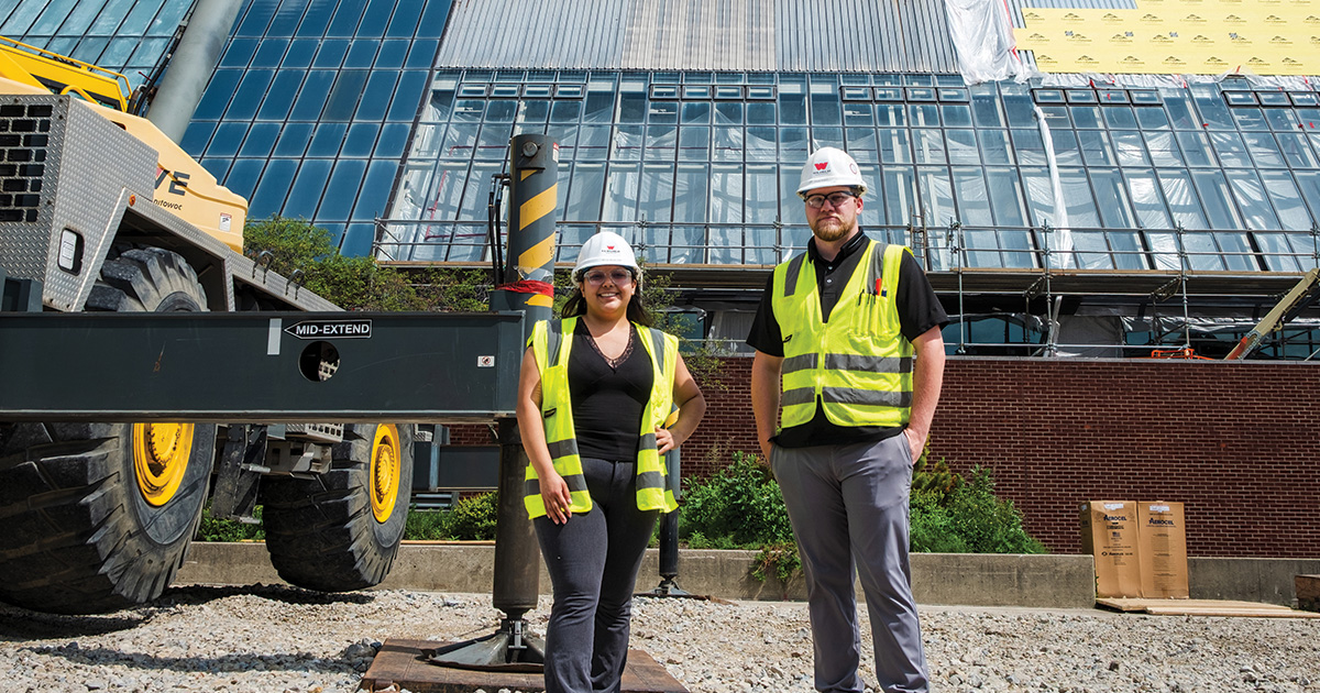 Two construction staff members wearing hard hats and reflective safety vests stand in front of a building under renovation, with construction equipment and scaffolding behind them.