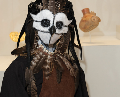 Elaborate owl-inspired costume made with feathers, fabric, and a white-and-black face covering, displayed in a museum gallery.