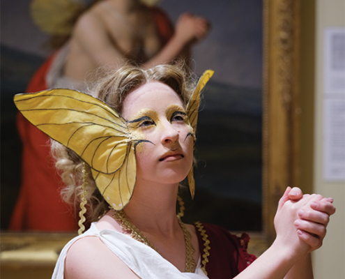 Performer wearing a fairy-inspired costume with large yellow wings and gold face paint poses in front of a framed painting in an art museum.