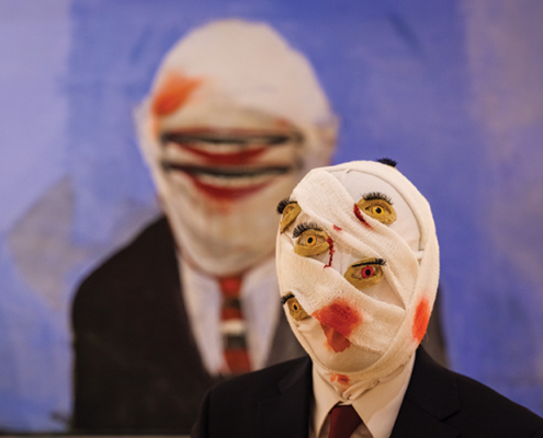 Performer in a dark suit wears a bandage-wrapped mask decorated with multiple painted eyes, posed in front of a large portrait.