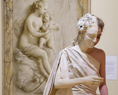 Performer in a white classical-style costume and face covering poses in front of a framed relief sculpture in an art museum.