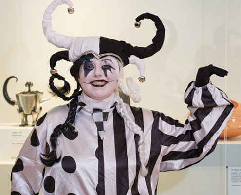 Performer in a black-and-white jester costume and horned hat poses in front of display cases in an art museum.