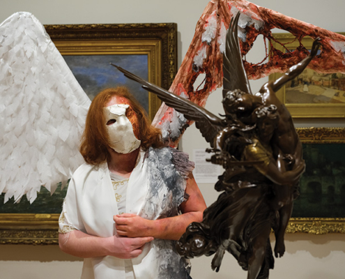 Performer wearing a white mask and large contrasting wings stands beside a bronze winged sculpture in an art museum.