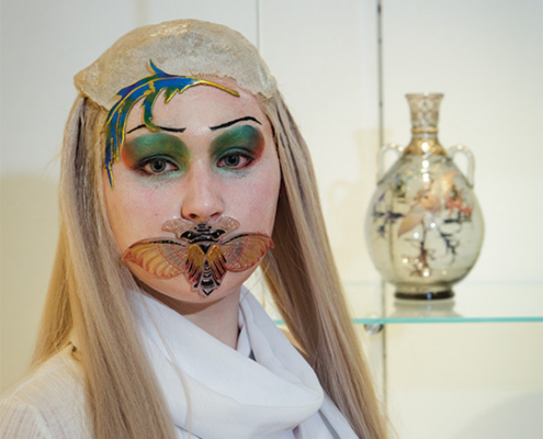 Performer with long blond hair and colorful face paint wears a decorative moth-like mouthpiece while standing in front of a ceramic vase in an art museum.
