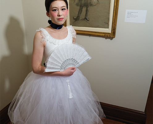 Performer in a white ballet-style dress holds a lace fan while posing beside a framed drawing of a similarly dressed figure in an art museum.