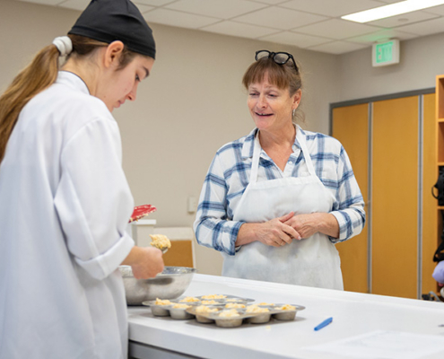 Student in a chef uniform prepares food at a counter while an instructor wearing an apron looks on.