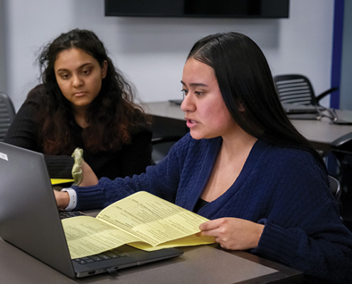 Two students work together at a table, reviewing documents and a laptop in a classroom.