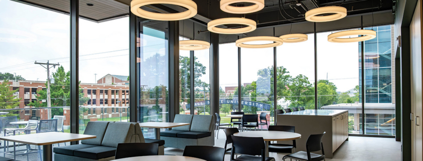 Modern lounge and collaboration space with tables, seating, large windows, and circular ceiling lights overlooking the Ball State campus.