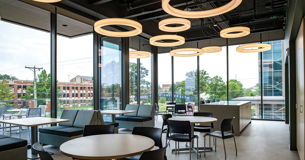 Modern lounge and collaboration space with tables, seating, large windows, and circular ceiling lights overlooking the Ball State campus.