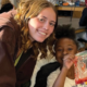 A woman stands. next to a girl in a classroom who inholding out a project they're working on.
