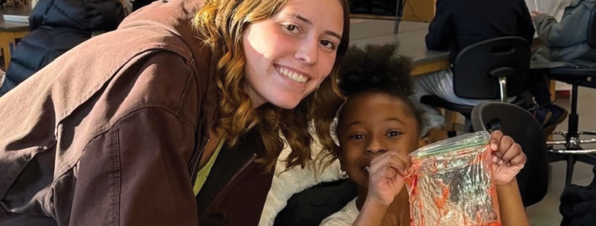 A woman stands. next to a girl in a classroom who inholding out a project they're working on.