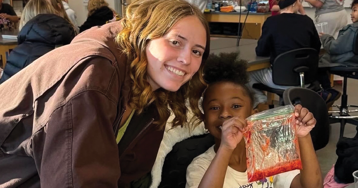 A woman stands. next to a girl in a classroom who inholding out a project they're working on.