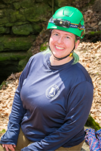 A woman sits outside smiling, wearing a brightly colored helmet for cave exploration.